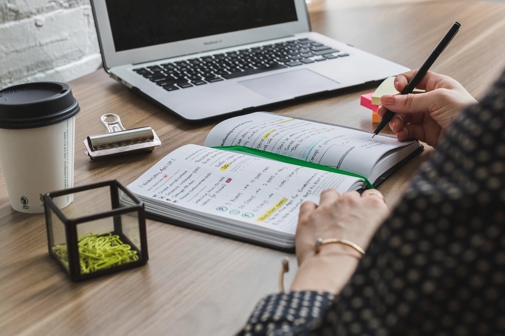 A person writing a to-do list in a notebook beside a laptop